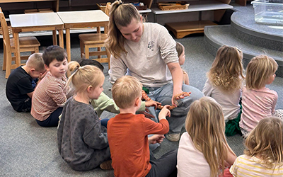 Staff member holding an orange and white cornsnake for children to look at. Children looking at the snake and gently touching it with two fingers.
