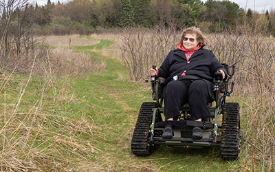 A winter prairie trail with a green path and dry brush in the background. A person traveling down the trail in an all-terrain wheelchair.