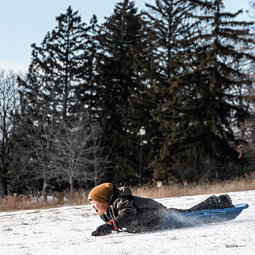 Child laying on their stomach on a sled going down the hill with a huge smile on their face.