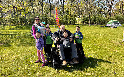 A group of people, including a person in a wheelchair, smiling outdoors on a grassy field with trees and a tent in the background